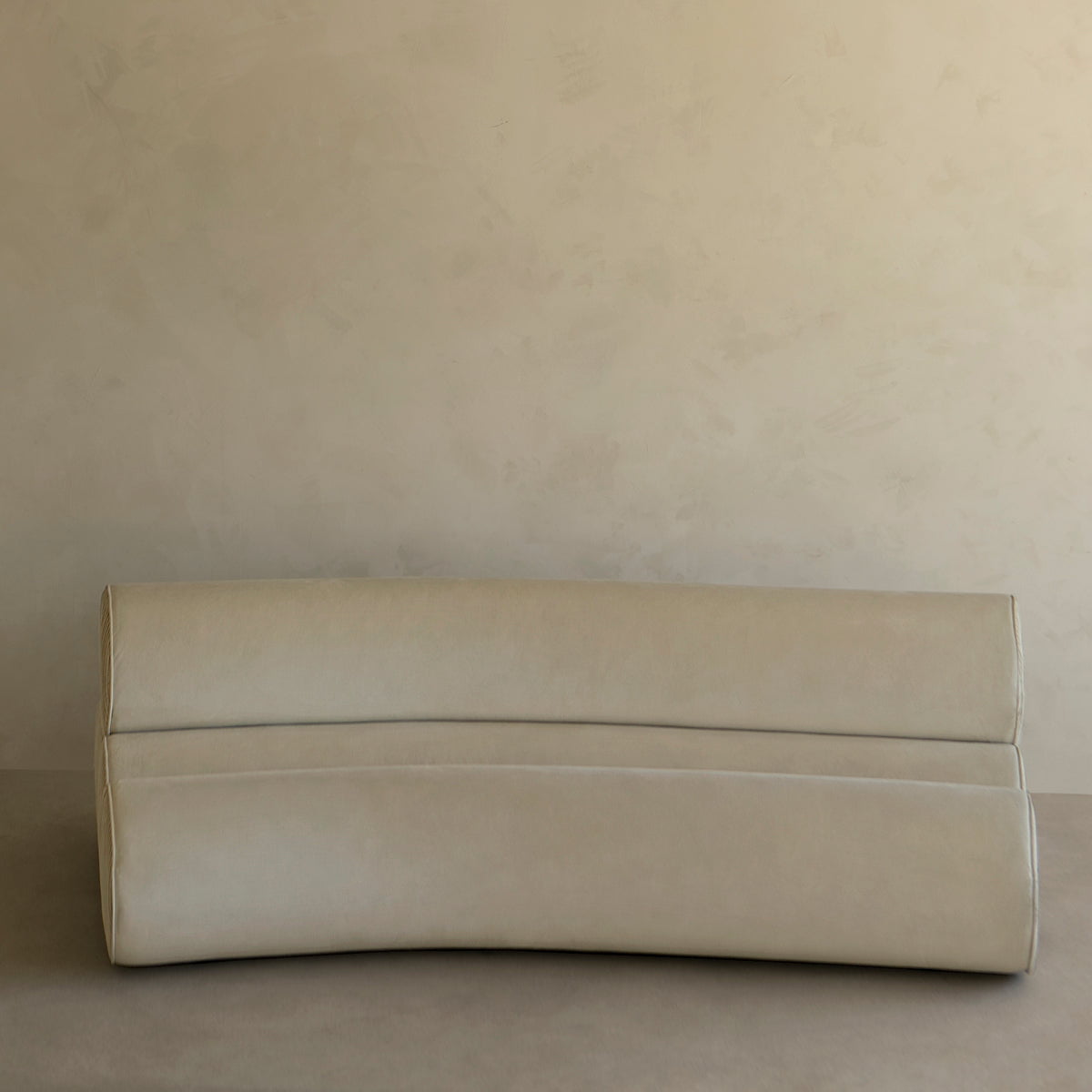 A minimalist, modern room showcases the Cylindra Sofa Rental—a light beige neotenic sofa with a curved backrest and base—set against matching beige walls and flooring in a simple, neutral-toned space.