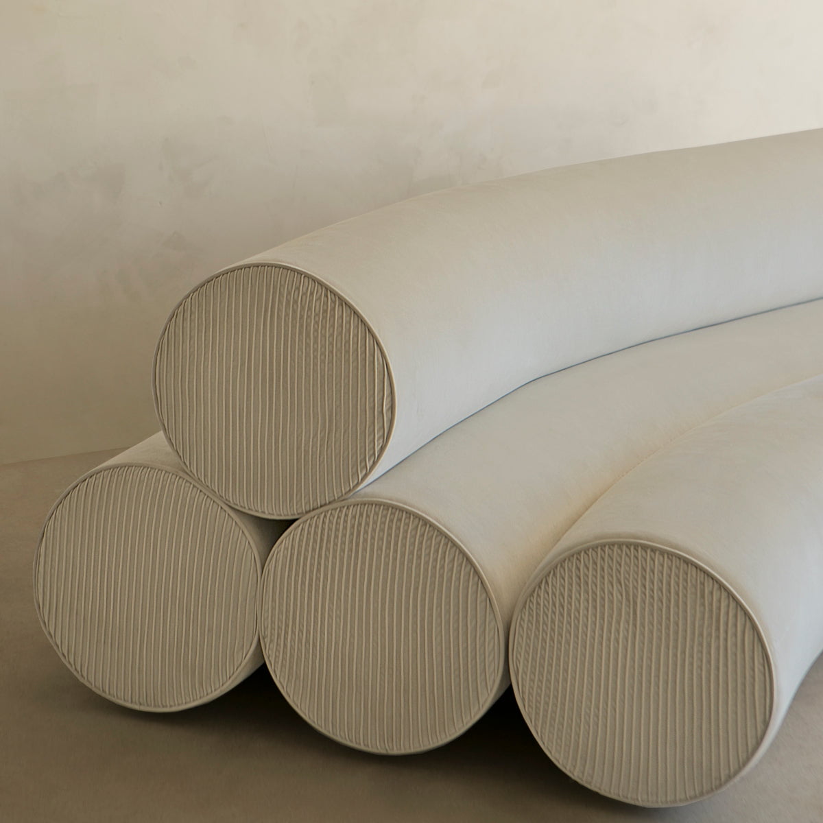 Four large, cream-colored Cylindra Sofa cushions—three stacked on the bottom and one on top—create a neotenic look. The cylindrical cushions have subtly striped ends and are set against a neutral, textured wall background.
