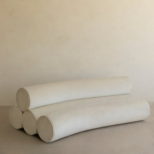 The Cylindra Sofa Rental features four large, cylindrical cushions in two layers and light beige upholstery, creating a minimalist look ideal for modern spaces. It sits elegantly against a plain beige wall for a sleek aesthetic.