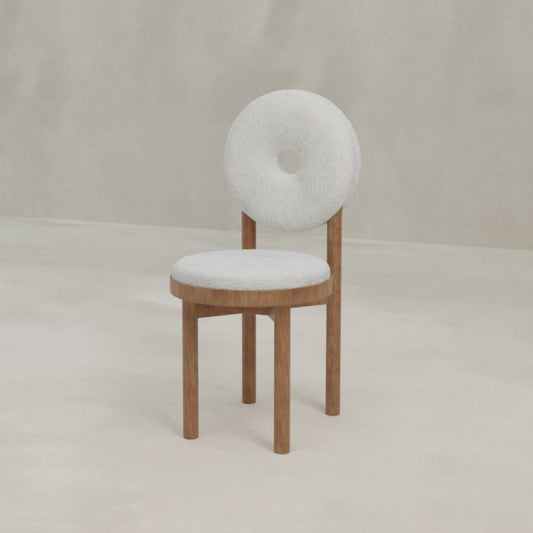 The Donut Chair features a modern design with a round, light gray cushioned seat and backrest—each with a central hole—and wooden legs, making it a striking statement piece against the plain light beige background.