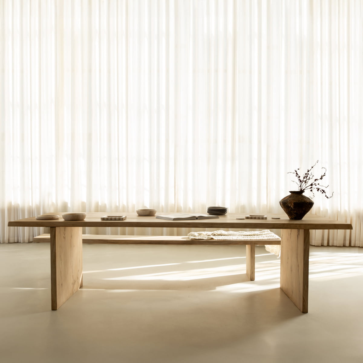 The Elm Farmhouse Table Large, featuring a minimalist silhouette and crafted from elm wood, is styled with pottery, books, and a woven cloth in a bright room with sheer white curtains and soft natural light.