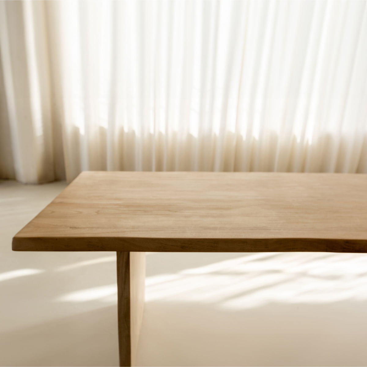 The Elm Farmhouse Table Large, featuring a minimalist silhouette in elm wood, stands on a light-colored floor bathed in soft sunlight through sheer white curtains.