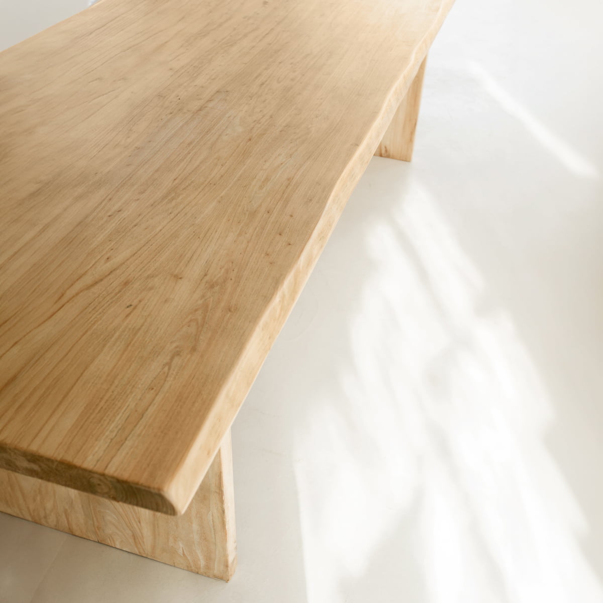 A close-up of the Elm Farmhouse Table Large, crafted from elm wood with a natural smooth finish, set on a light floor with soft sunlight and gentle shadows nearby.