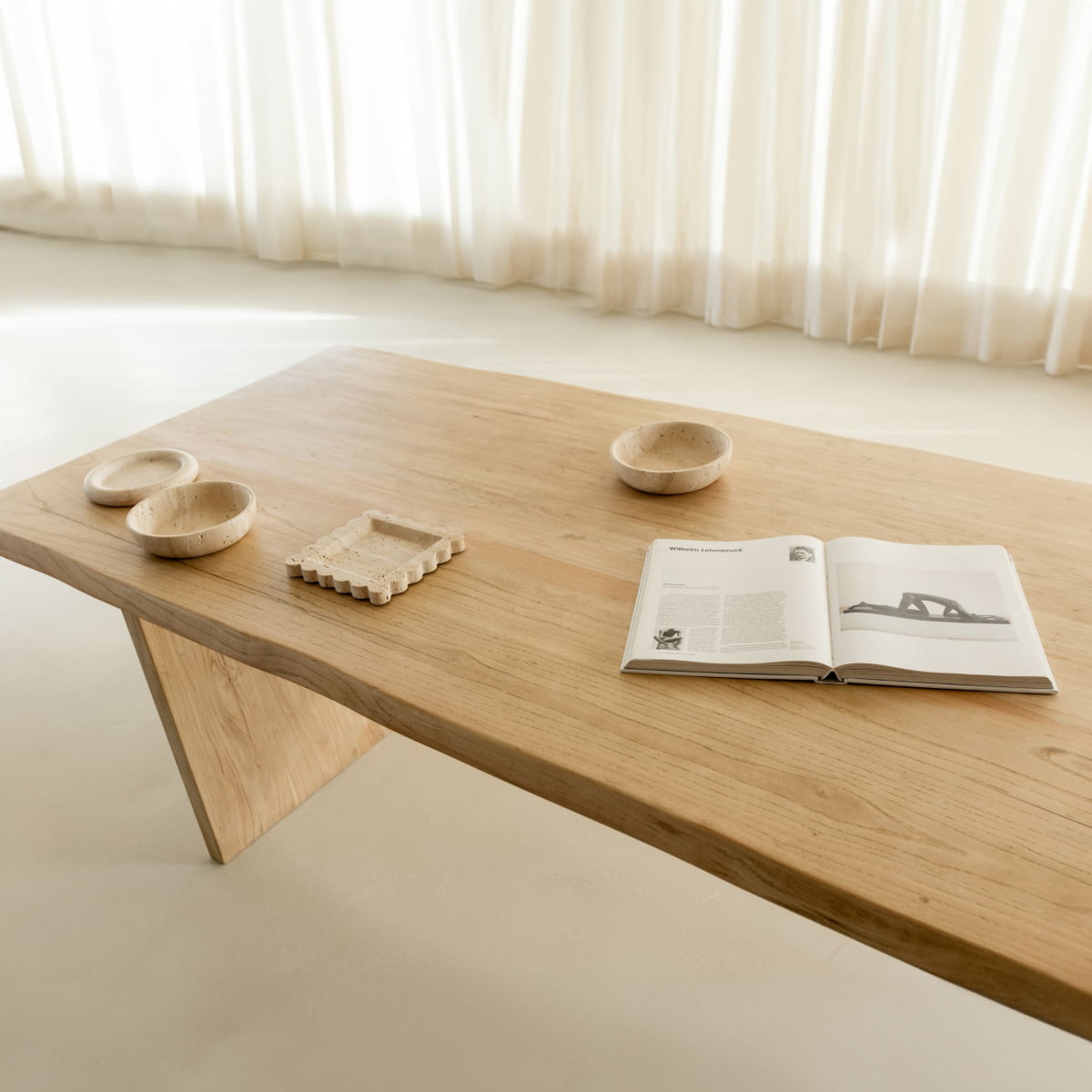 The Elm Farmhouse Table Large features a minimalist wooden design, styled with three light-toned bowls, a decorative tray, and an open book, set in a bright room with sheer white curtains in the background.