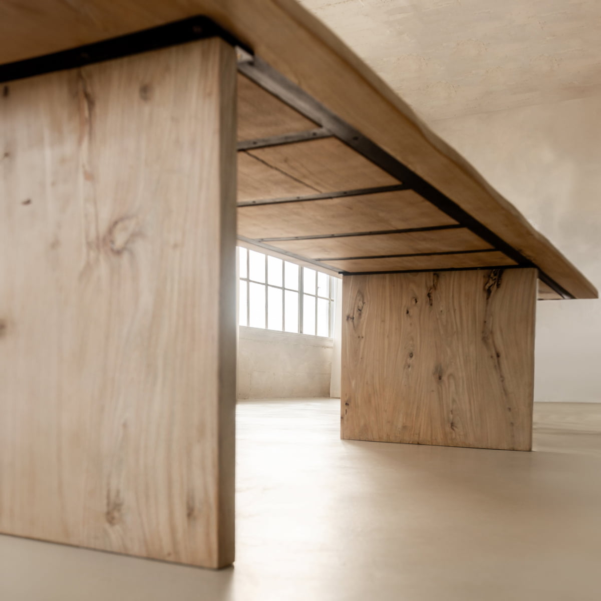 Low-angle view of the Elm Farmhouse Table Large, featuring thick legs and a natural finish, set in a bright, minimalistic room with light flooring and a large window letting in plenty of natural light.