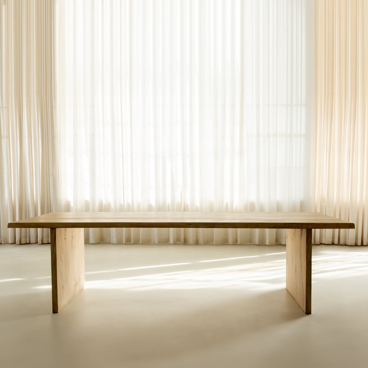 A minimalist silhouette defines the Elm Farmhouse Table Large, crafted from elm wood and featured in a bright room with sheer white curtains filtering natural light in the background.