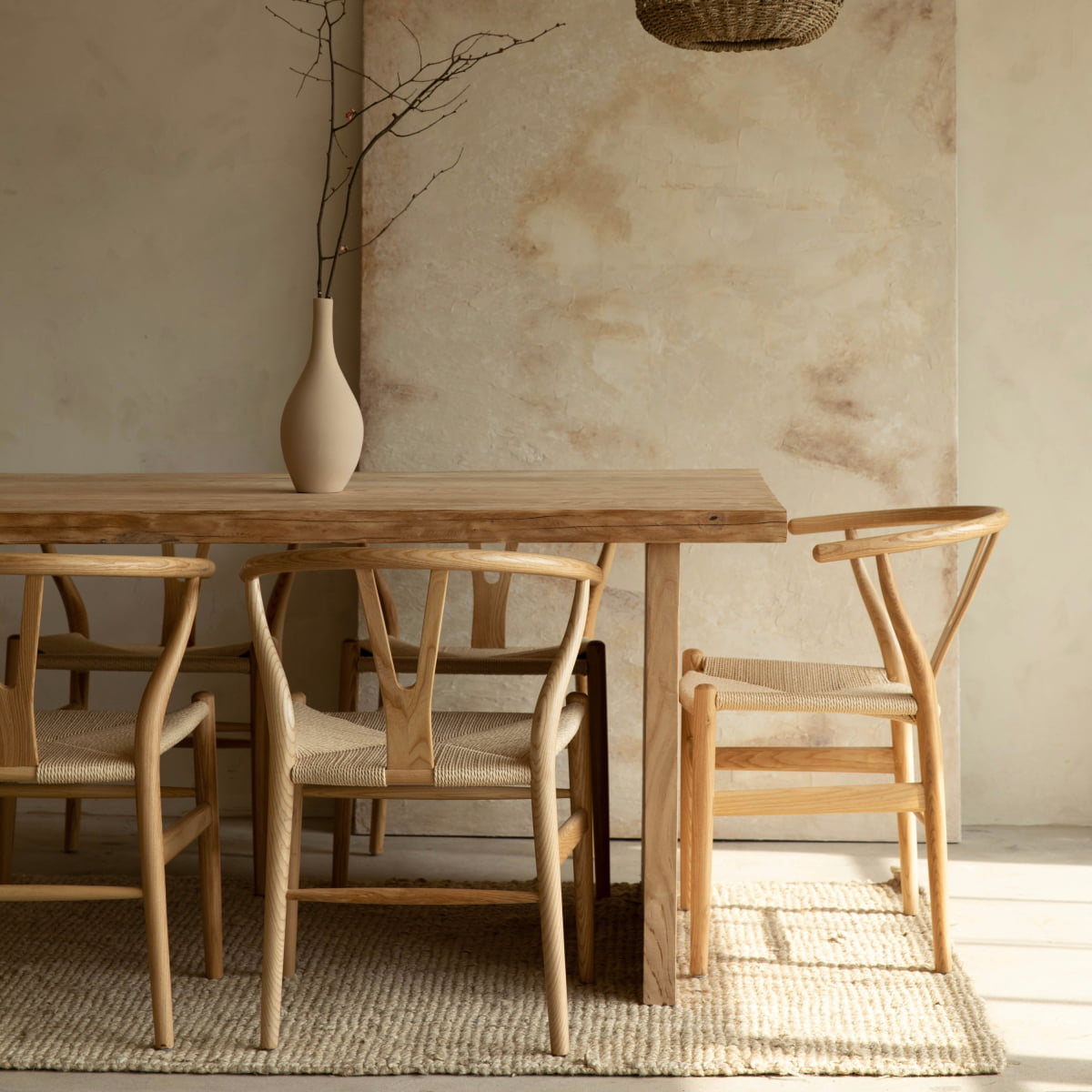 A minimalist dining room features a farmhouse dining table, four Elsa Chairs with woven seats and Y-back design, a beige rug, and a tall vase with branches on the table, set against a textured, neutral-toned wall.