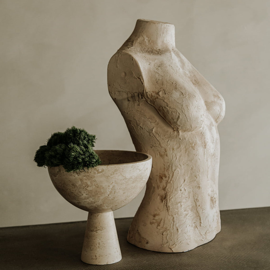 A textured beige female torso sculpture sits next to the Elysian Pedestal Bowl, a travertine pedestal bowl with green foliage, both displayed on a gray surface against a neutral background.