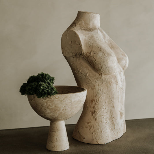 A textured beige female torso sculpture sits next to the Elysian Pedestal Bowl, a travertine pedestal bowl with green foliage, both displayed on a gray surface against a neutral background.