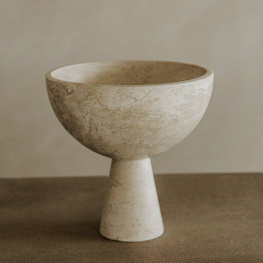 The Elysian Pedestal Bowl is a minimalist, round travertine piece with a textured surface and conical pedestal, displayed on a neutral surface against a soft beige background—perfect for refined home decor.