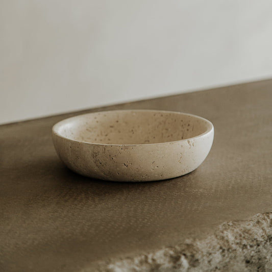 The Étoile Bowl in Medium, a round, shallow beige travertine piece, sits on a textured brown surface with a softly blurred neutral background.