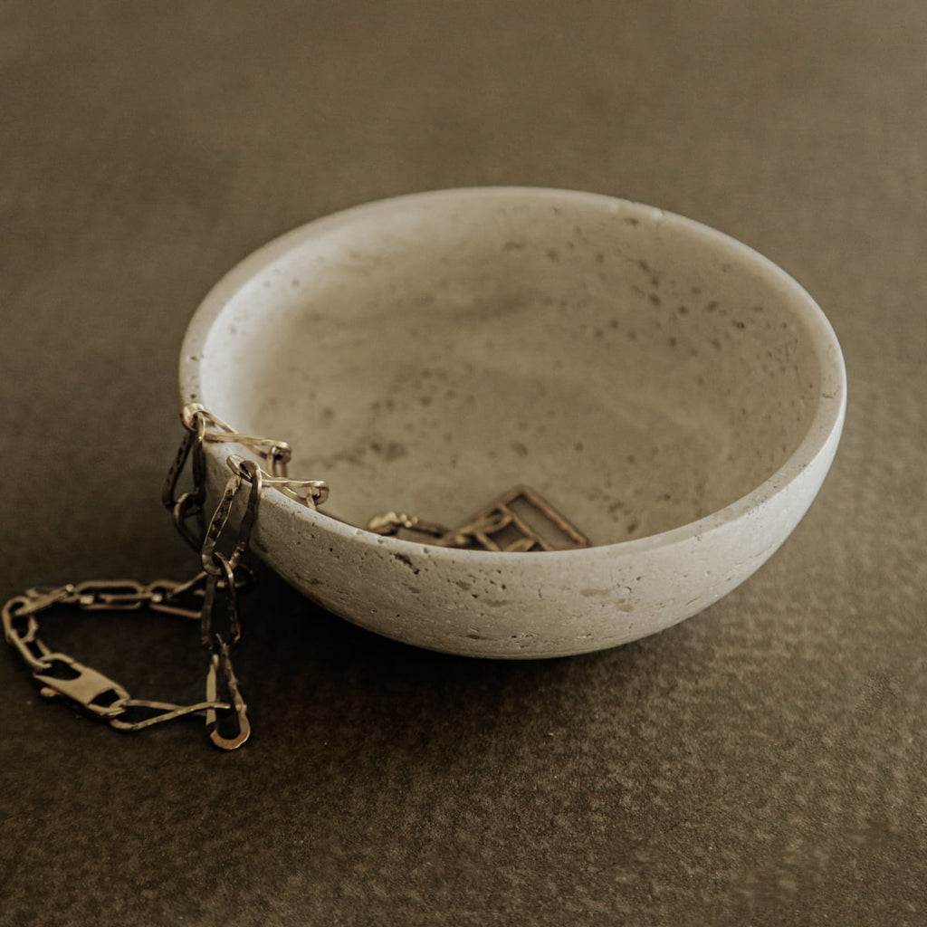 The Étoile Bowl in Small, crafted from concrete with French elegance, displays a gold chain draped inside and over its edge, set atop a textured brown surface.