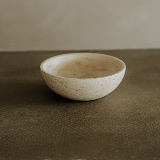 A small, round Étoile Bowl in light beige travertine rests on a textured brown surface against a neutral background, embodying French elegance and timeless style.