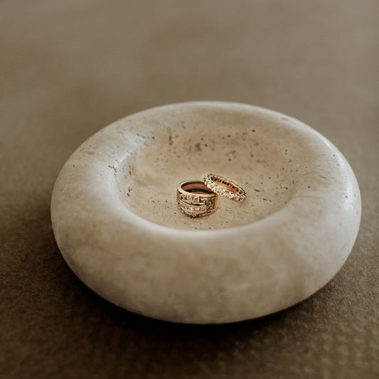 Two gold rings, one adorned with diamonds, rest in the center of the Étoile Bubble Tray—a shallow, round travertine piece—on a brown surface, bringing a touch of French elegance to any space.