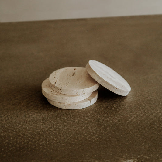 Four round, beige Étoile Coasters are stacked on a textured brown surface, with one upright for display, showcasing French elegance and premium travertine craftsmanship in the Étoile Coaster Set of 4.