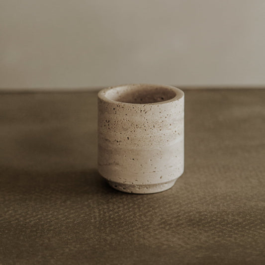 The Étoile Travertine Pen Holder, a small cylindrical beige accessory with a speckled finish, adds French elegance to any desk as it rests on a brown textured surface against a simple background.