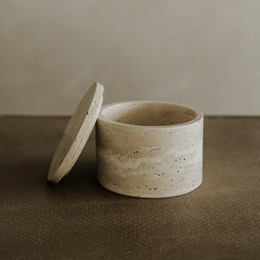 The Étoile Vault with lid, a round beige travertine organizer, sits on a textured brown surface, bringing French elegance to the neutral background.
