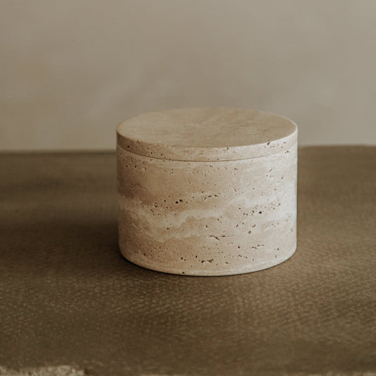 The Étoile Vault with lid is a round, beige travertine organizer featuring a smooth finish and natural stone patterns, showcased on a textured brown surface with a neutral background. Its multifunctional design suits various uses.