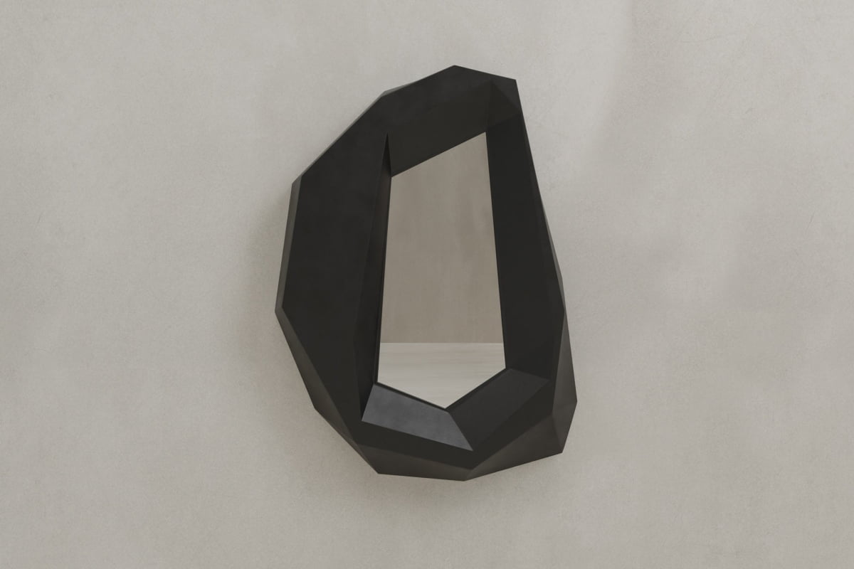 The Facet Mirror Large features a geometric, irregular shape with a thick black angular frame. Mounted on a light gray wall, its brutalist design and sculptural form offer modern sophistication reminiscent of a faceted gemstone.