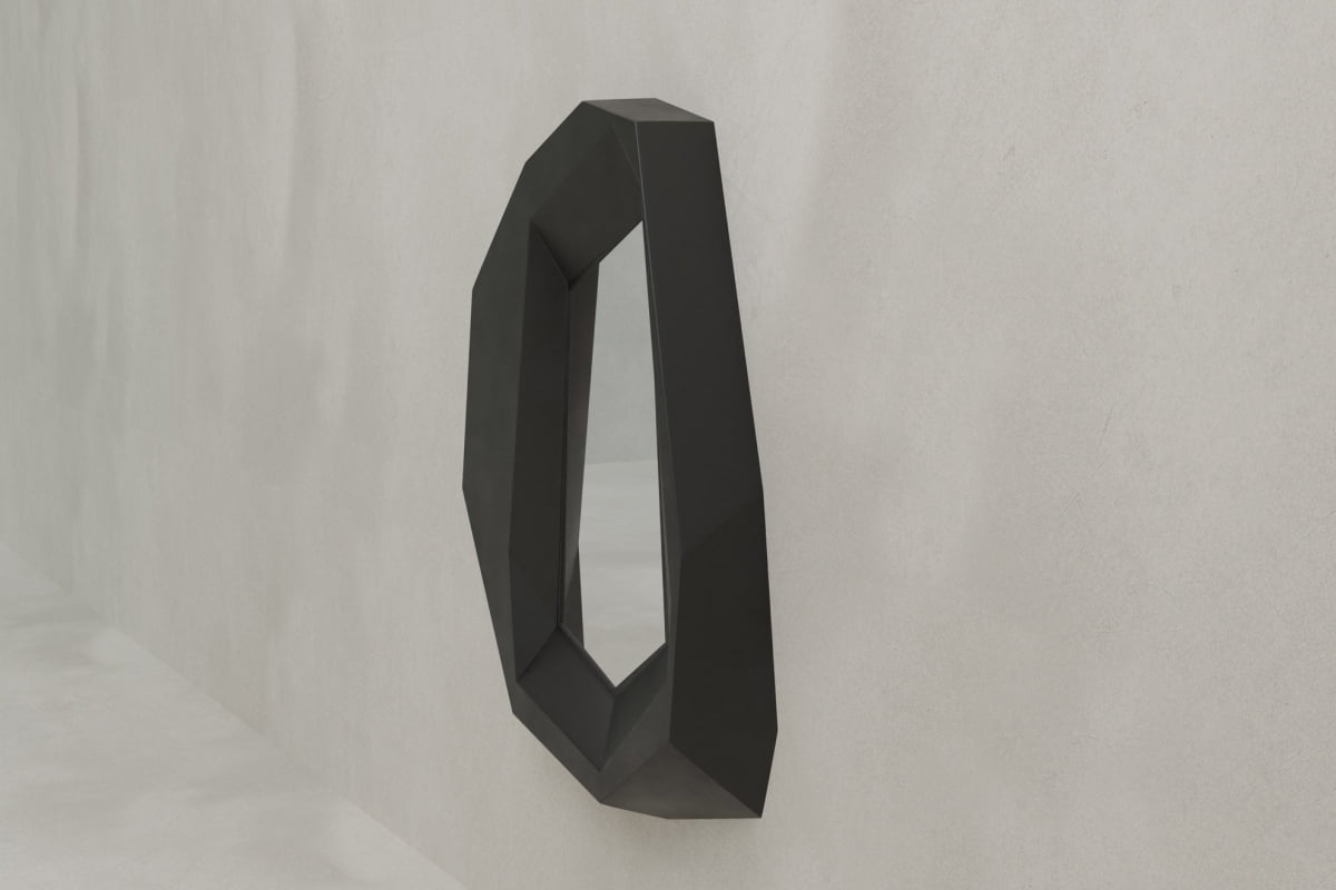 The Facet Mirror Large, featuring a black frame and geometric, irregular angles, is mounted on a light gray wall, bringing modern sophistication and a bold, minimalist brutalist design to the space.