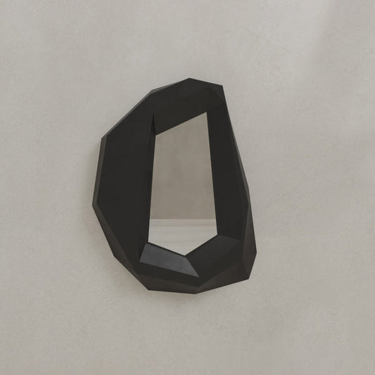 The Facet Mirror Small, with its angular black frame and irregular geometric shape, hangs on a light wall, adding modern sophistication to the minimalist space.