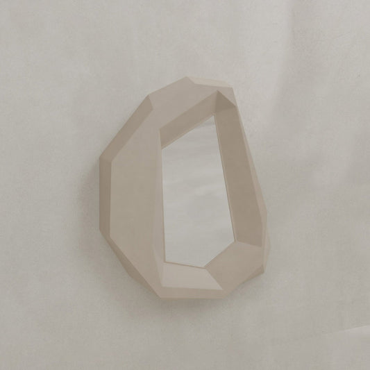 The Facet Mirror Small features a beige, angular frame and an irregular shape inspired by brutalist design, resembling a cut crystal. Hung on a light-textured wall, it adds modern sophistication and style to any room.