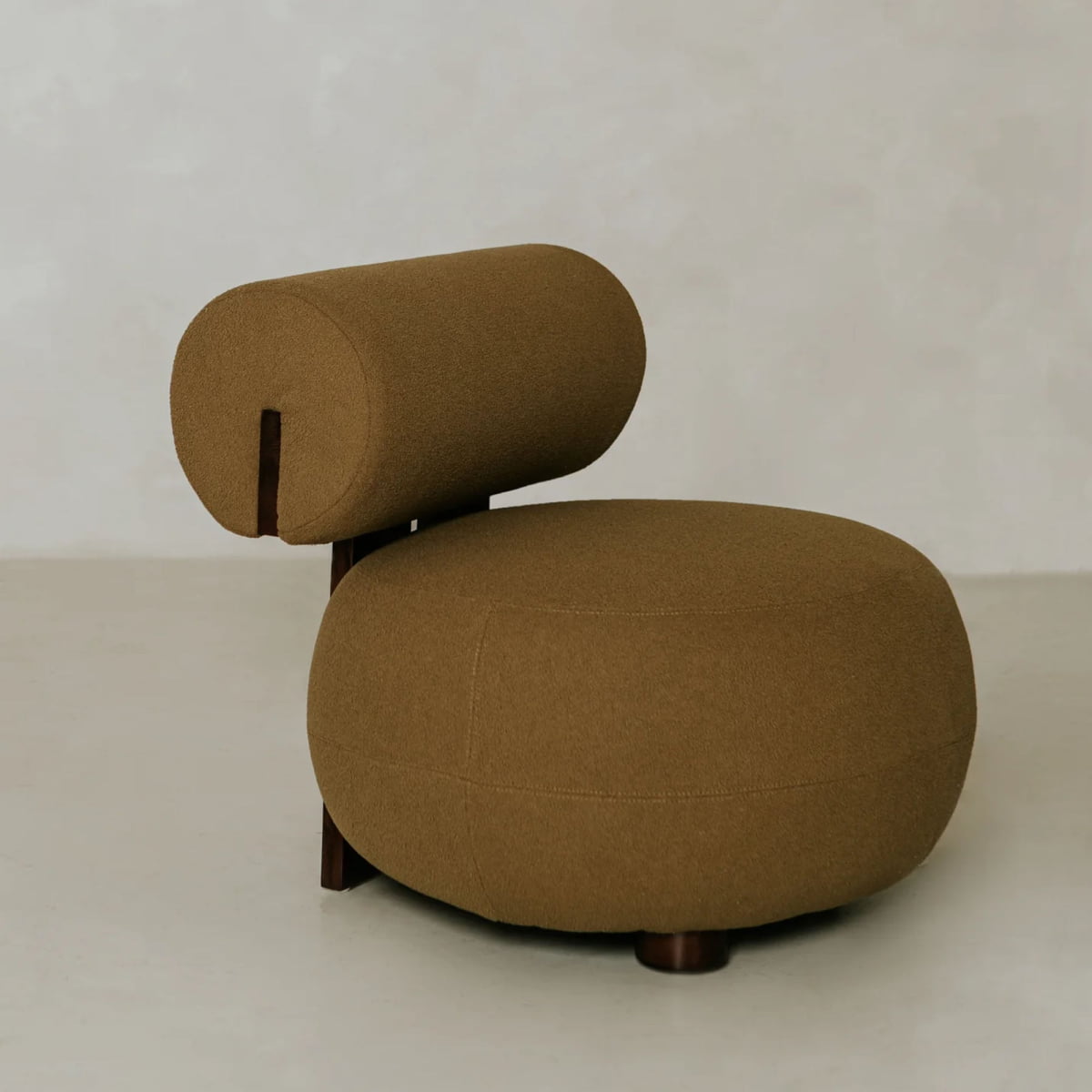 The Finchley Chair is a modern, minimalist lounge chair featuring a round brown cushioned seat, a matching cylindrical backrest, and solid wooden legs that emphasize its sleek wooden profile against a light neutral backdrop.