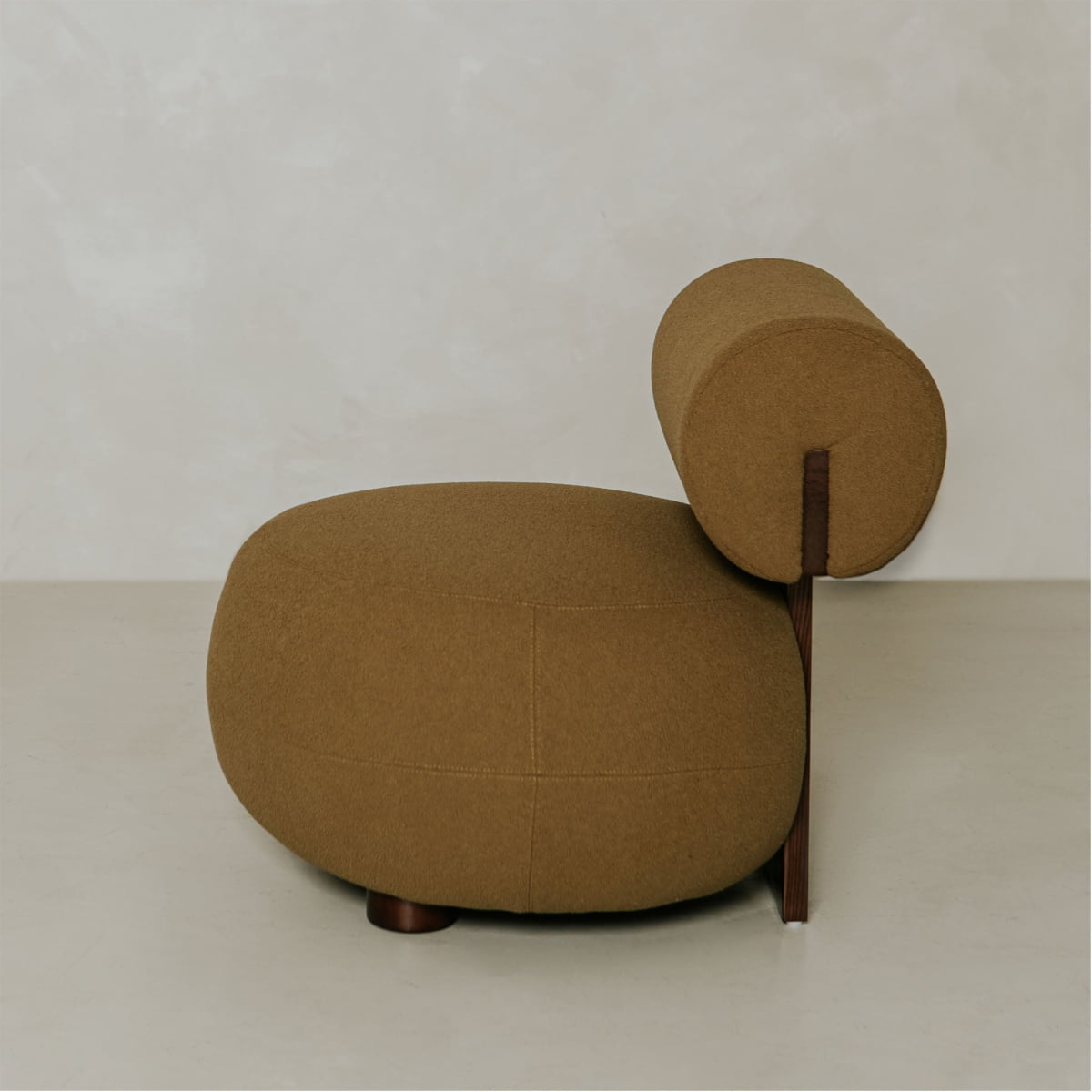 The Finchley Chair is a modern, minimalist brown lounge chair with a rounded seat, cylindrical backrest, sleek wooden profile, and solid legs, shown against a light neutral background.
