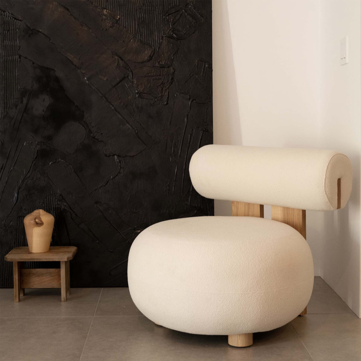 A modern, minimalist room features the round Finchley Chair with solid legs beside a small wooden table holding a beige hand-shaped sculpture, all set against a textured black wall panel.