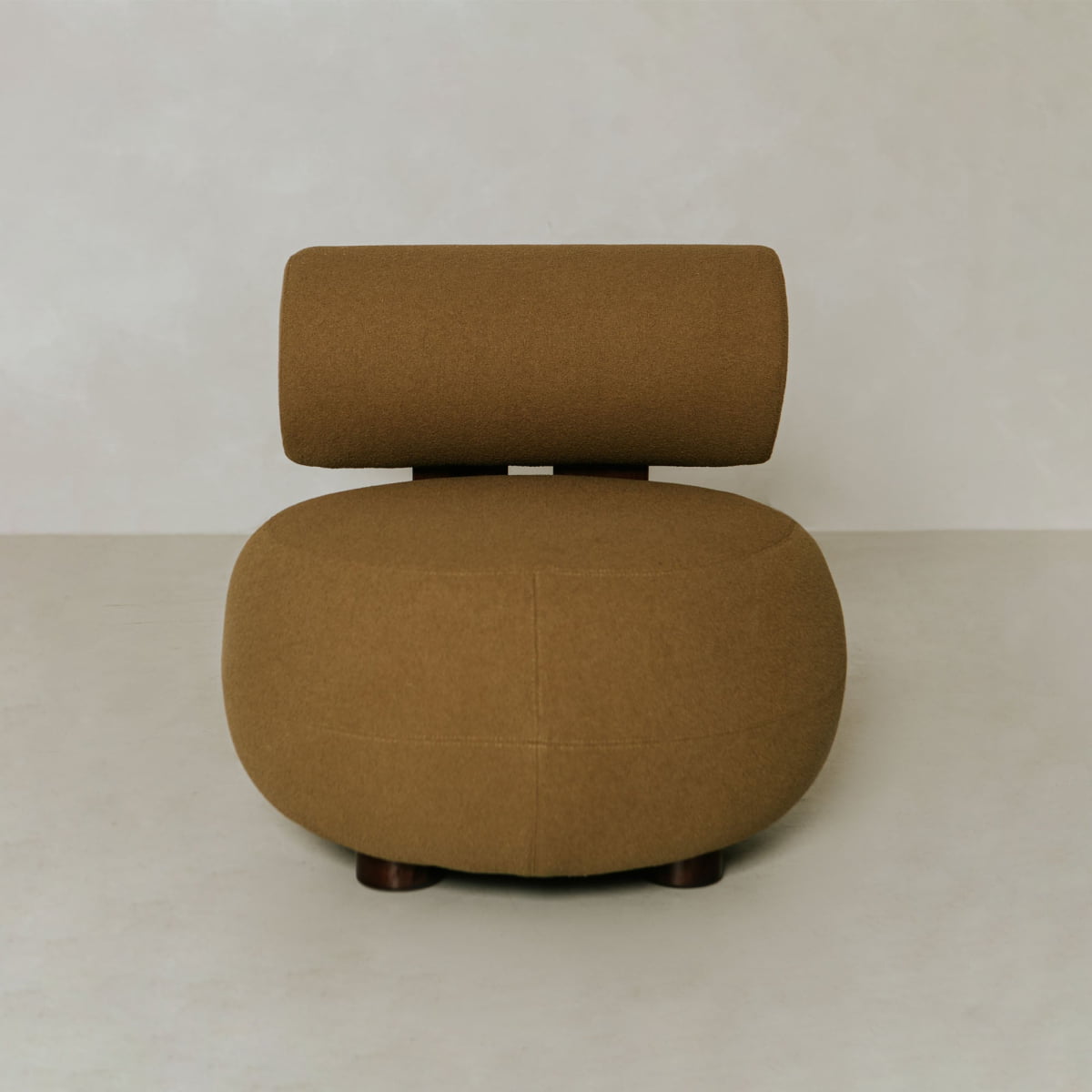 The Finchley Chair features a modern, minimalist wooden profile with a rounded brown upholstered seat and matching cylindrical backrest, standing on solid legs atop a light-colored floor against a neutral wall.