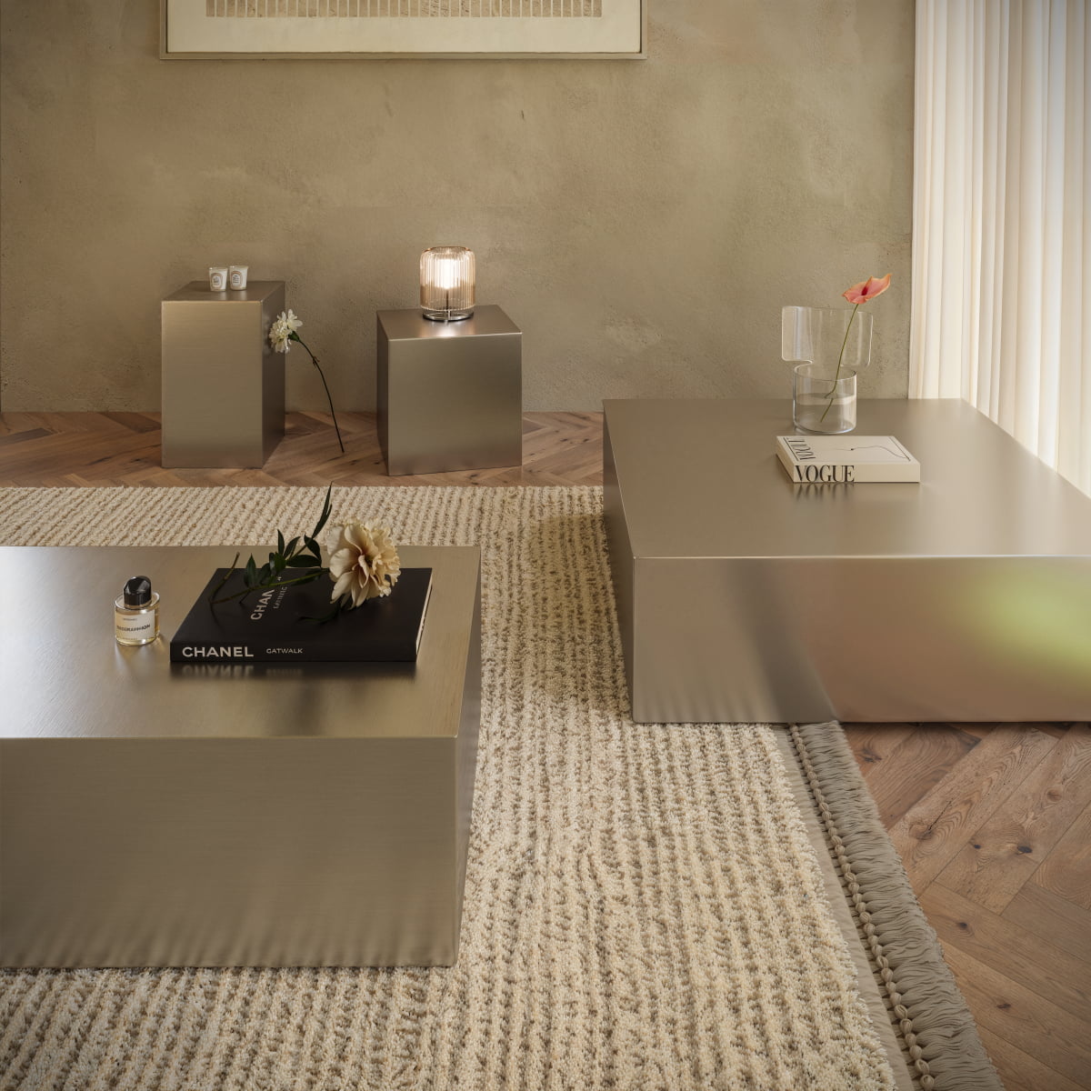 A minimalist living room with a neutral palette features the Forge Steel Tall Plinth as a metallic table, a textured beige rug, and decorative items. Soft natural light highlights the modern design and subtle industrial vibe of the space.