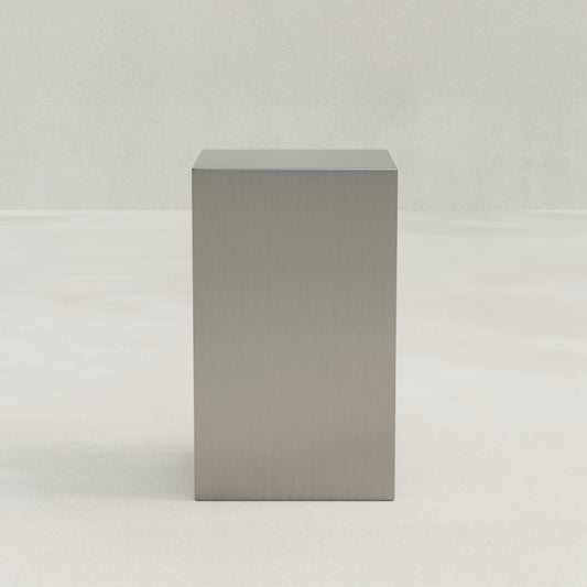 The Forge Steel Tall Plinth stands on a smooth, light-colored surface against a pale background, adding modern design and subtle industrial flair to the space.