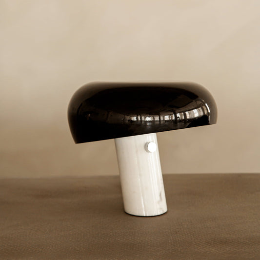 The Freya Lamp features a modern, minimalist design with a glossy black dome-shaped shade and a slanted white cylindrical marble base, beautifully set against a neutral beige background.