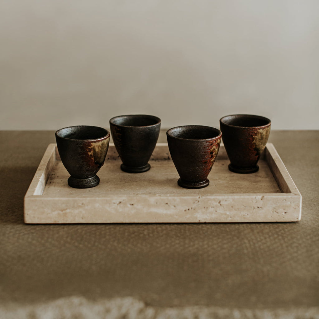 Four small, dark ceramic cups sit on the Grande Cascade Tea Tray, their elegant forms contrasting beautifully with the tray’s textured surface and neutral background.