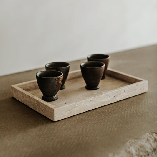 The Grande Cascade Tea Tray, a rectangular piece crafted from travertine, displays four small dark ceramic cups arranged elegantly on its textured brown surface against a neutral backdrop.