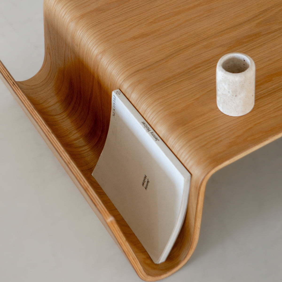 A close-up of the Gregory Coffee Table features smooth, curved wooden edges. A white book fits neatly in a built-in groove, while a cylindrical stone cup sits on top—ideal for enhancing any living space.