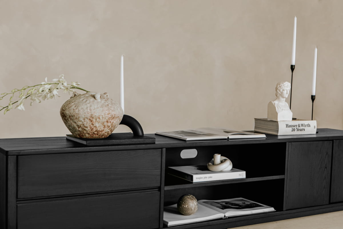 The Indre Media Console offers ample storage and a sleek modern look, styled with a rustic vase of white flowers, tall candles, stacked books, a bust sculpture, and open magazines against a beige wall for refined minimalism.