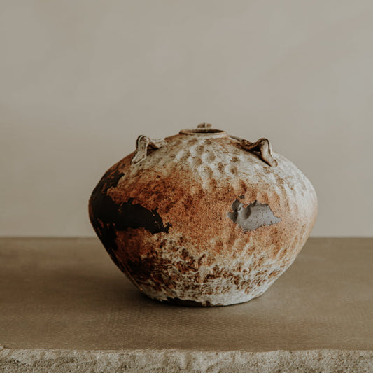 Ishi is a rustic ceramic pot in earthy brown and white tones, featuring an uneven, weathered finish and small protrusions near the top. Ideal for home decor, it sits beautifully on any surface against a plain background.