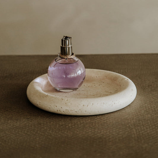 A round glass perfume bottle with purple liquid rests on the Janvry Tray, its circular Travertine design enhancing style and function atop a textured brown surface against a plain light background.