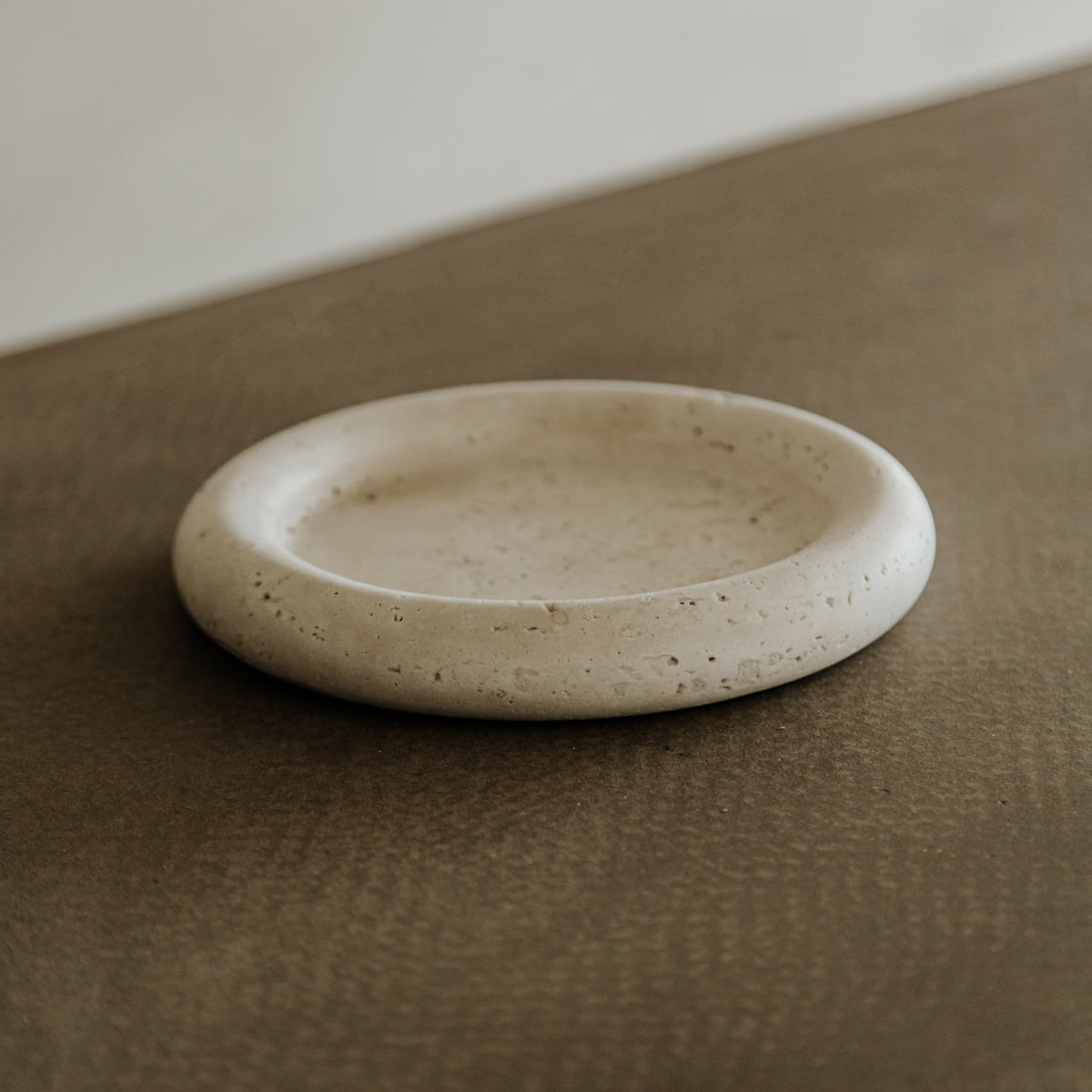 The Janvry Tray, a round, shallow travertine piece with a subtly textured surface, rests on a brown tabletop and effortlessly combines natural style with practical everyday use.