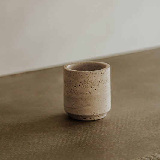 Le Chalice, a small beige ceramic cup with a speckled texture, rests on a travertine-style brown surface against a softly blurred backdrop, bringing sophisticated elegance to the dining setting.