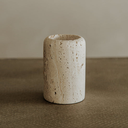 Le Gobelet, a cylindrical beige travertine vase with a rough, textured surface, rests on a smooth brown surface against a plain neutral background.