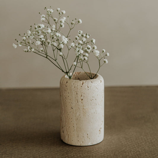 The Le Gobelet, a small round travertine vase, displays delicate white babys breath flowers against a neutral textured backdrop—an elegant accent for any refined dining setting.