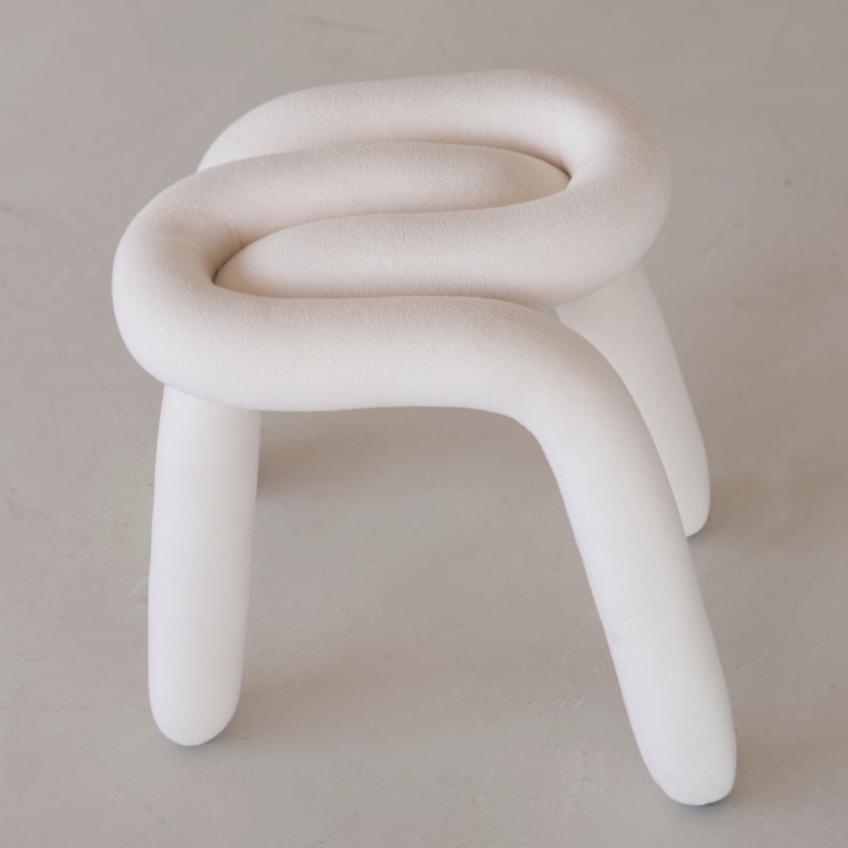 The Loud Stool features a sculptural modern design with three thick, curved white legs and a plush, looped top. Crafted from luxurious polyurethane foam, its velvety surface adds comfort while echoing tubular steel chair style.