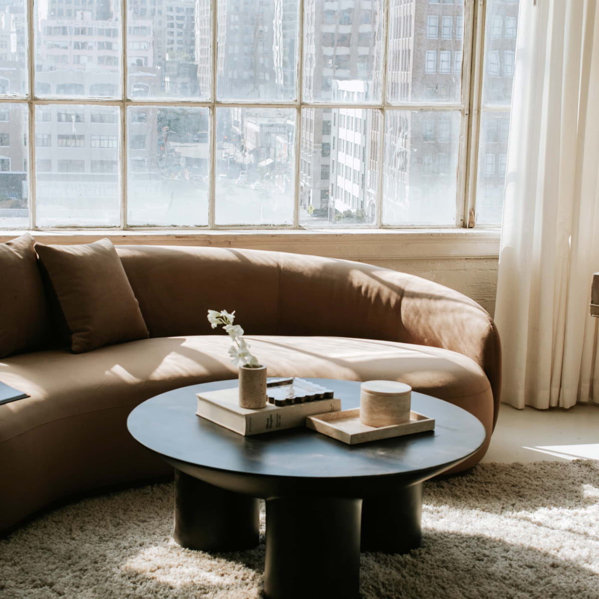 A modern living room with a curved brown sofa, the Luna Coffee Table featuring tubular legs, books and a small vase on a beige rug, all framed by large windows overlooking the cityscape—a true interior design statement.