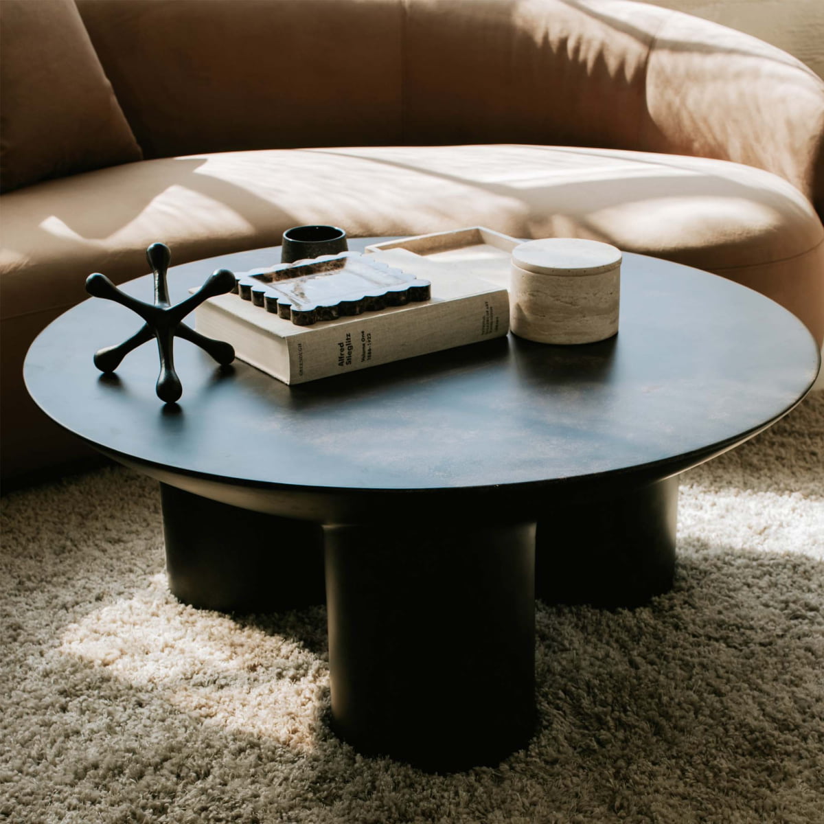 The Luna Coffee Table with thick tubular legs rests on a beige carpet in front of a tan curved sofa, highlighting chic interior design. On the table are a wooden tray, book, candle, and black jack sculpture as decor accents.