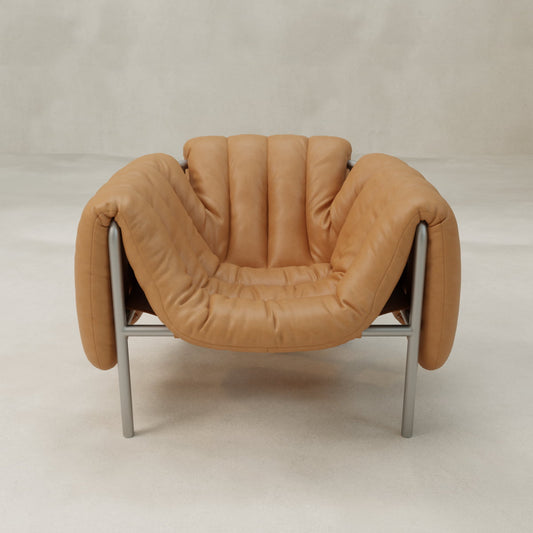 The Mallow Chair features soft tan leather upholstery with deep channeled tufting and plush cushioning, all supported by a sculptural gray metal tubular frame, creating a modern look against a neutral background.