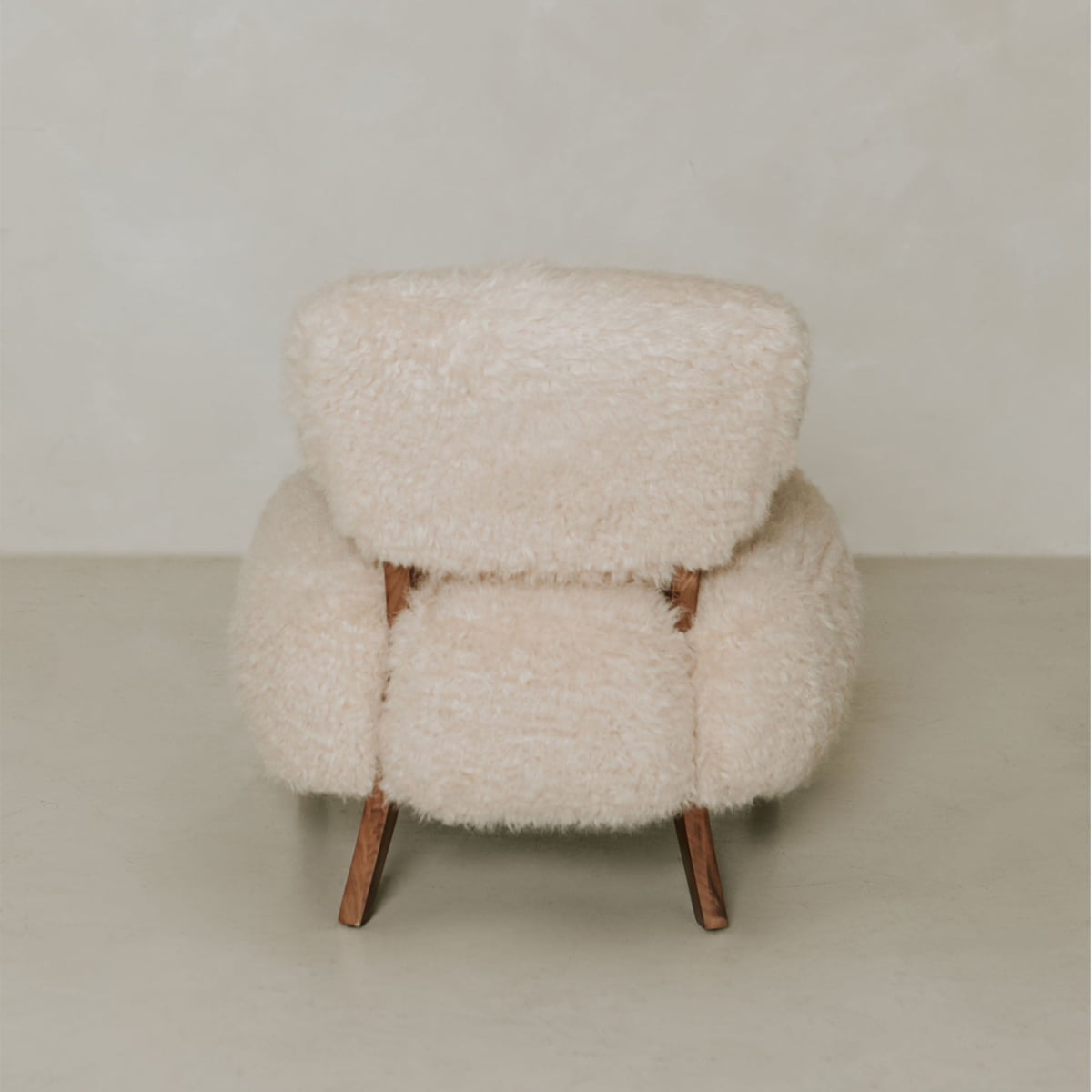 The Malmo Chair features thick, fluffy shearling upholstery and wooden legs, creating minimalist sophistication when placed against a plain light-colored wall and floor.