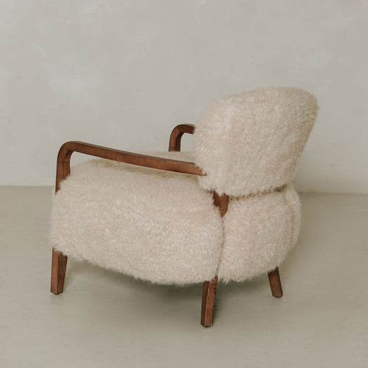 The Malmo Chair features wooden armrests and legs with plush cream shearling upholstery, offering minimalist sophistication against a plain, light background.