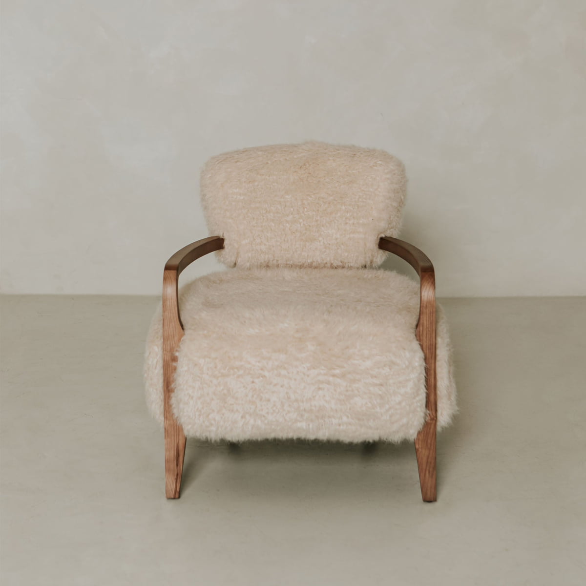 The Malmo Chair has wooden arms and legs and is upholstered in soft cream-colored shearling, creating a minimalist, sophisticated look against a plain, light-toned background.